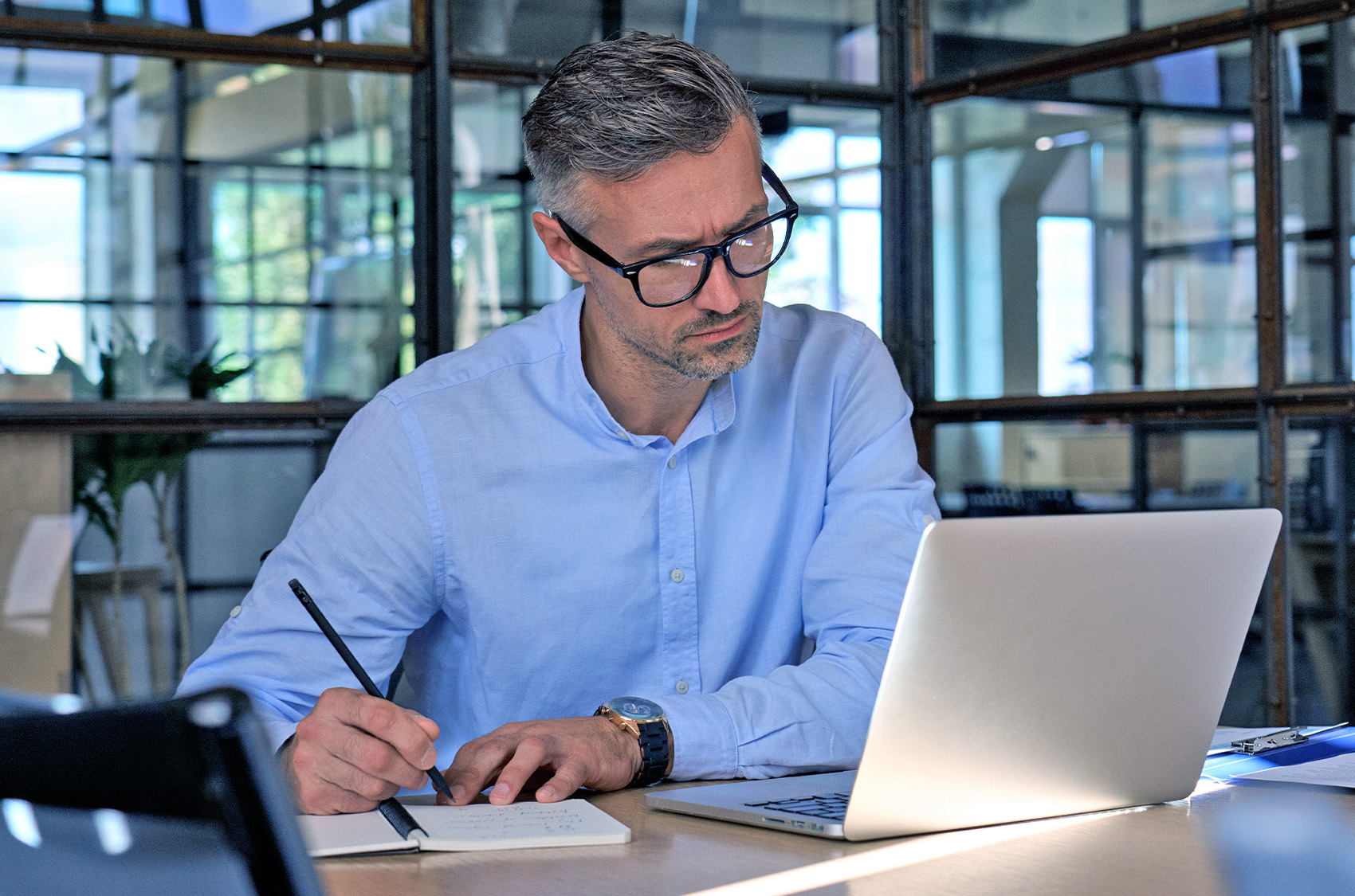 A man is sitting at a desk, looking at a notebook and holding a pen in his hand to take notes. Office